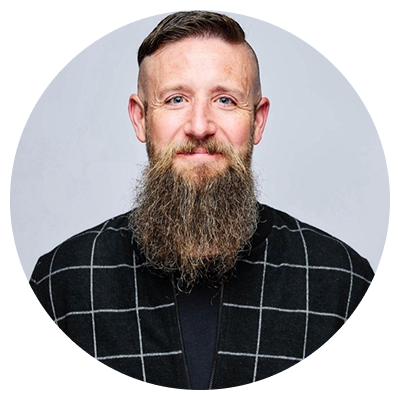Man with short hair and a long full beard wearing a black jacket with white grid pattern on light gray background.