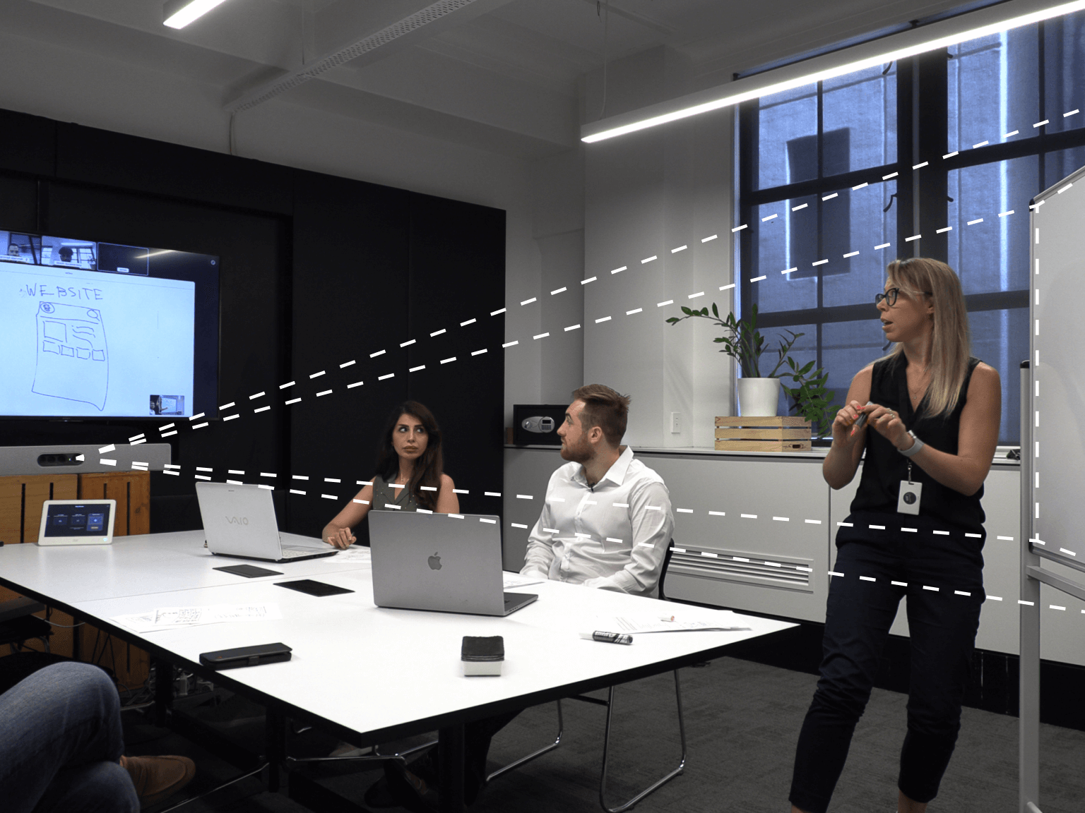 Three people in a modern office meeting room with laptops, a whiteboard, a large screen displaying a website sketch, and a woman standing and presenting.