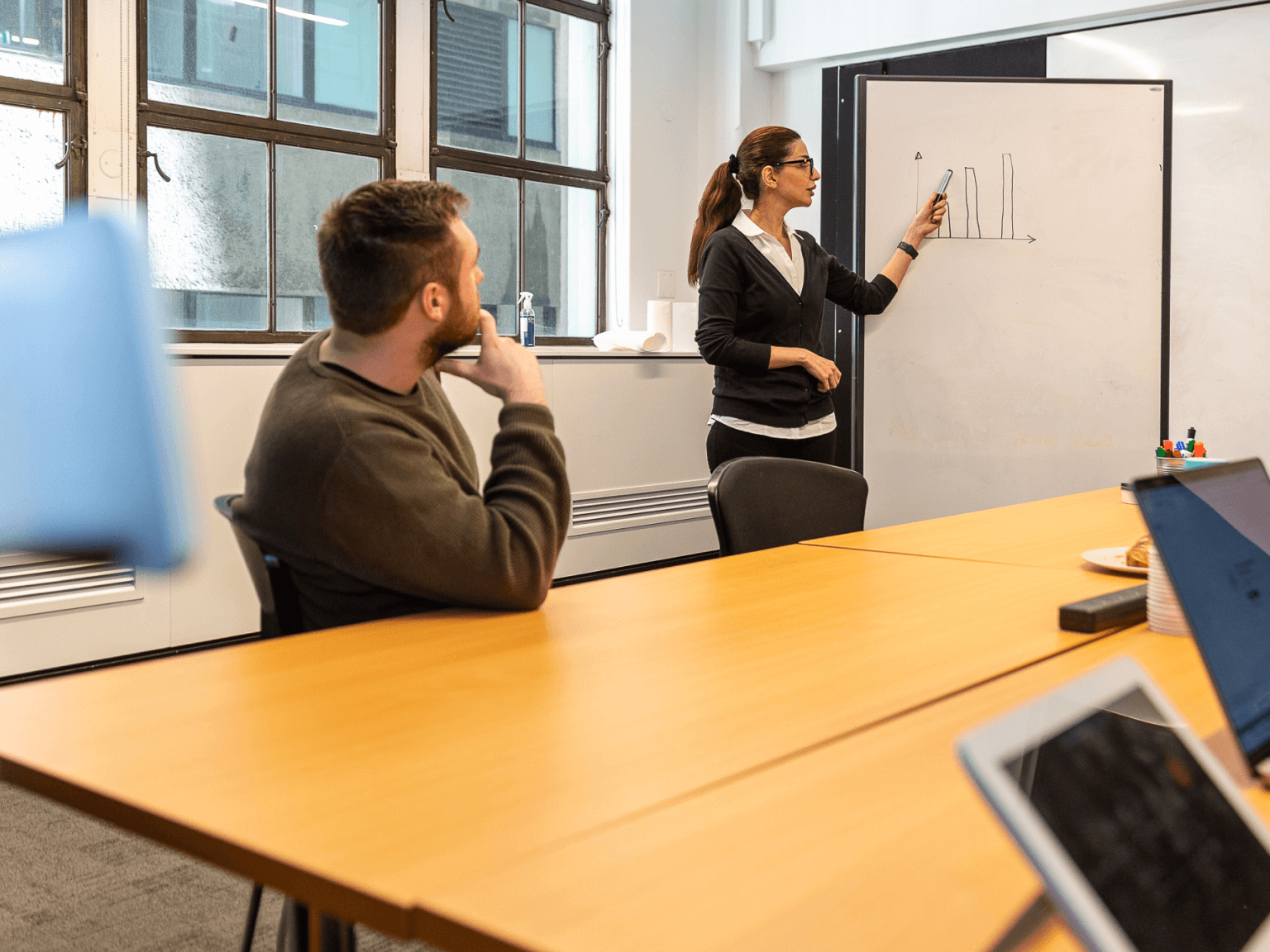 Woman presenting a bar chart on a whiteboard to a seated man in a modern conference room.