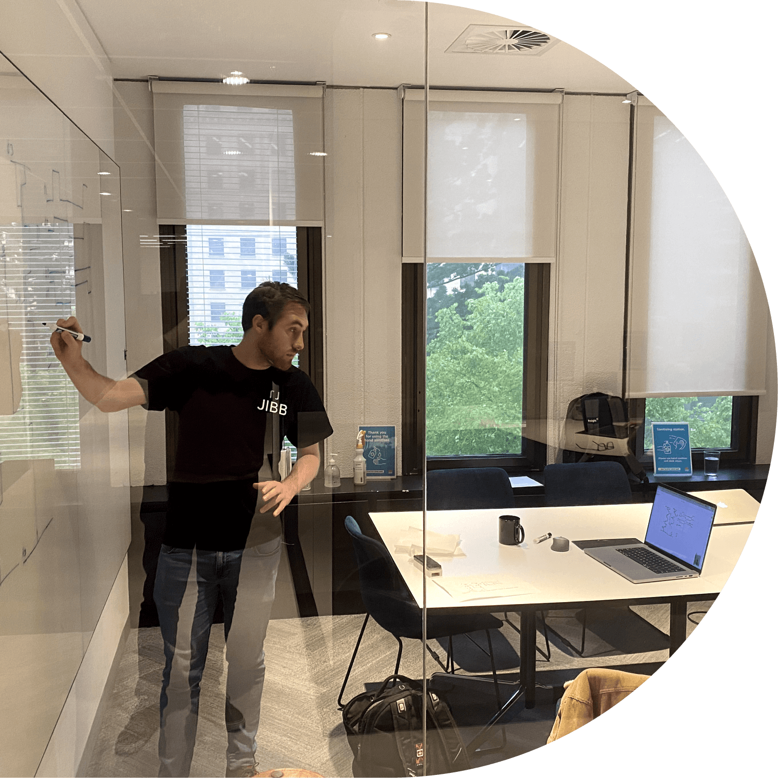 Man wearing a black JIBB shirt writing on a whiteboard inside a glass-walled office with a table, laptop, and chairs visible.