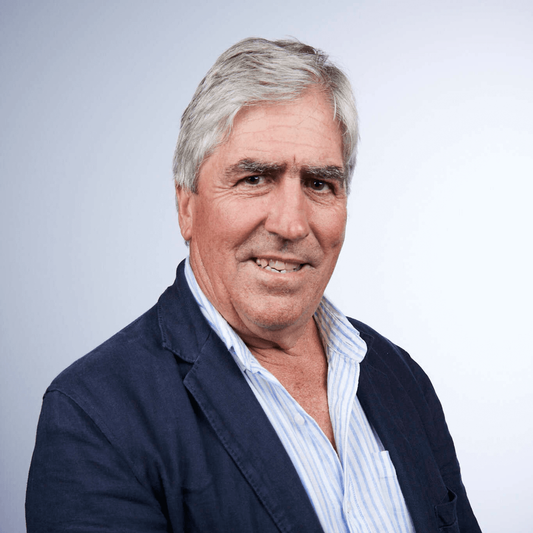 Older man with gray hair wearing a navy blazer over a blue and white striped button-up shirt, smiling slightly against a light gray background.
