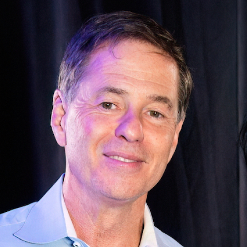 Middle-aged man with short brown hair wearing a light blue collared shirt, smiling against a dark background.
