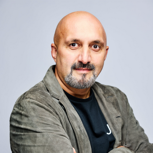 Bald man with a beard and mustache wearing a grey jacket and black shirt, posing with arms crossed against a light background.