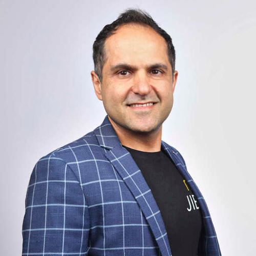 Smiling man with short dark hair wearing a blue checkered blazer over a black shirt against a light background.