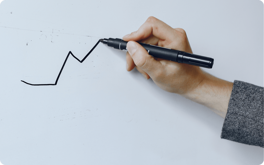 Hand holding a black marker drawing an upward trending zigzag line on a whiteboard.