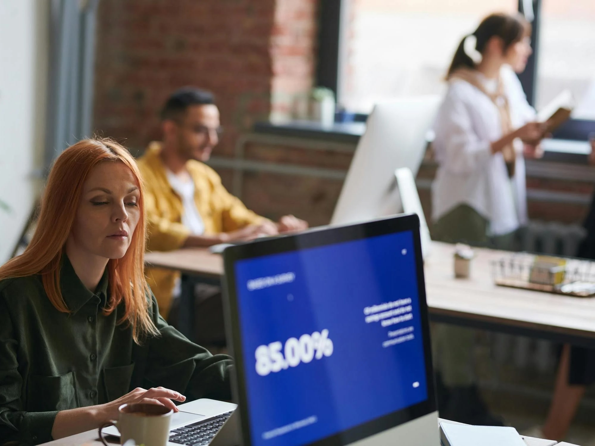 Woman working at a desk on a laptop next to a monitor displaying 85.00%, with two blurred coworkers in the background.