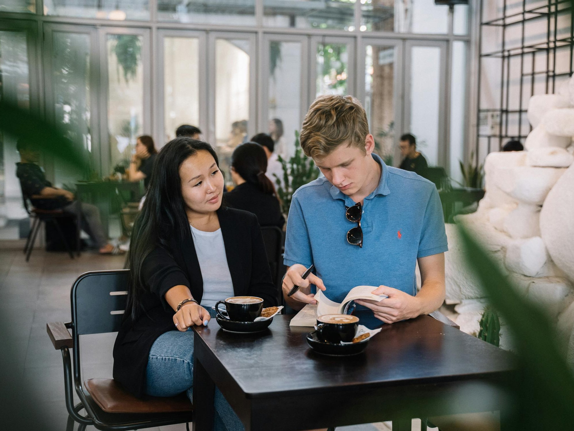 A man in a blue polo shirt reading a book and a woman in a black blazer sitting together at a table with two cups of coffee in a café.