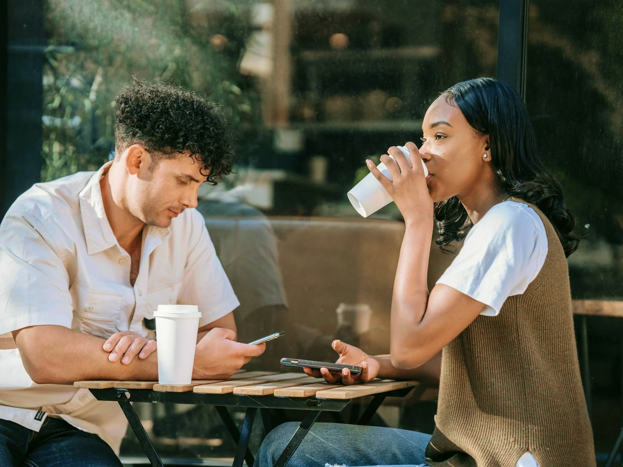 Man and woman sitting at a wooden table in a cafe, both holding smartphones, with the woman drinking from a white cup.