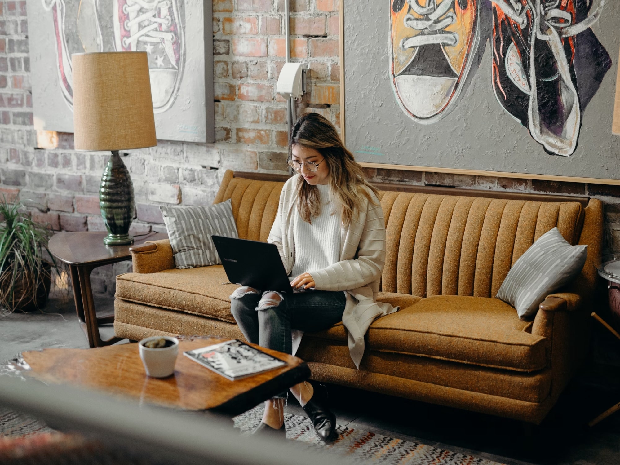 Woman with glasses sitting on a mustard yellow couch using a laptop in a room with brick walls and sneaker artwork.